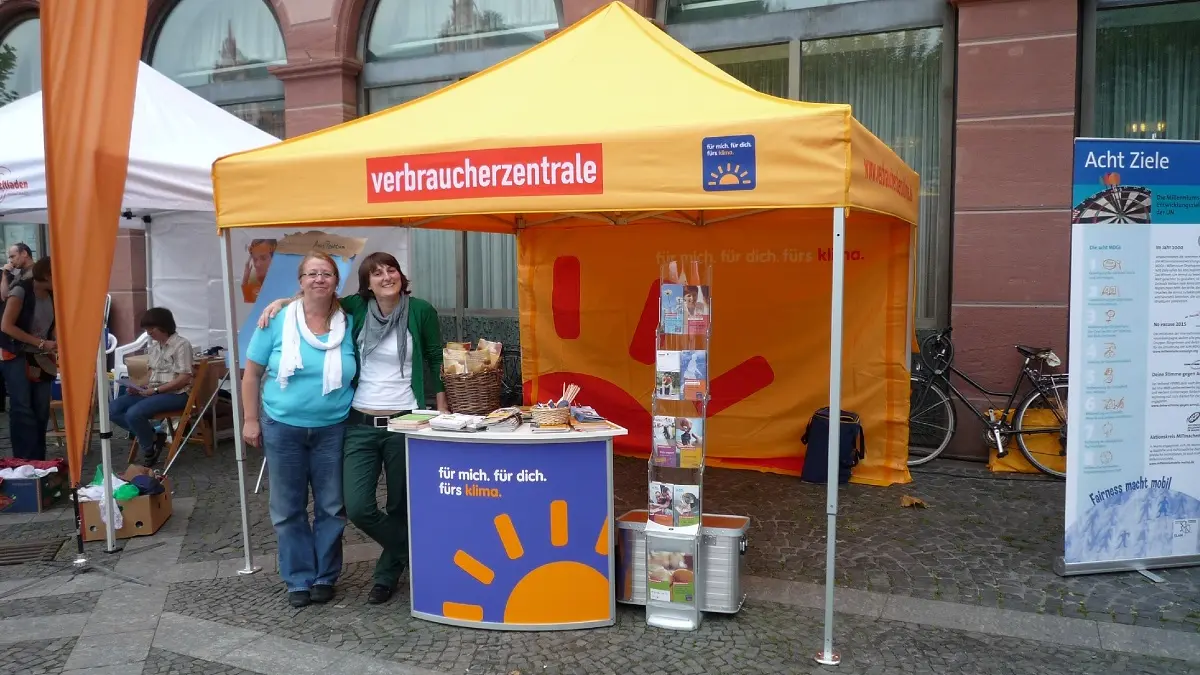 Zwei Frauen stehen an einem Straßenstand mit Infomaterial und blicken in die Kamera.
