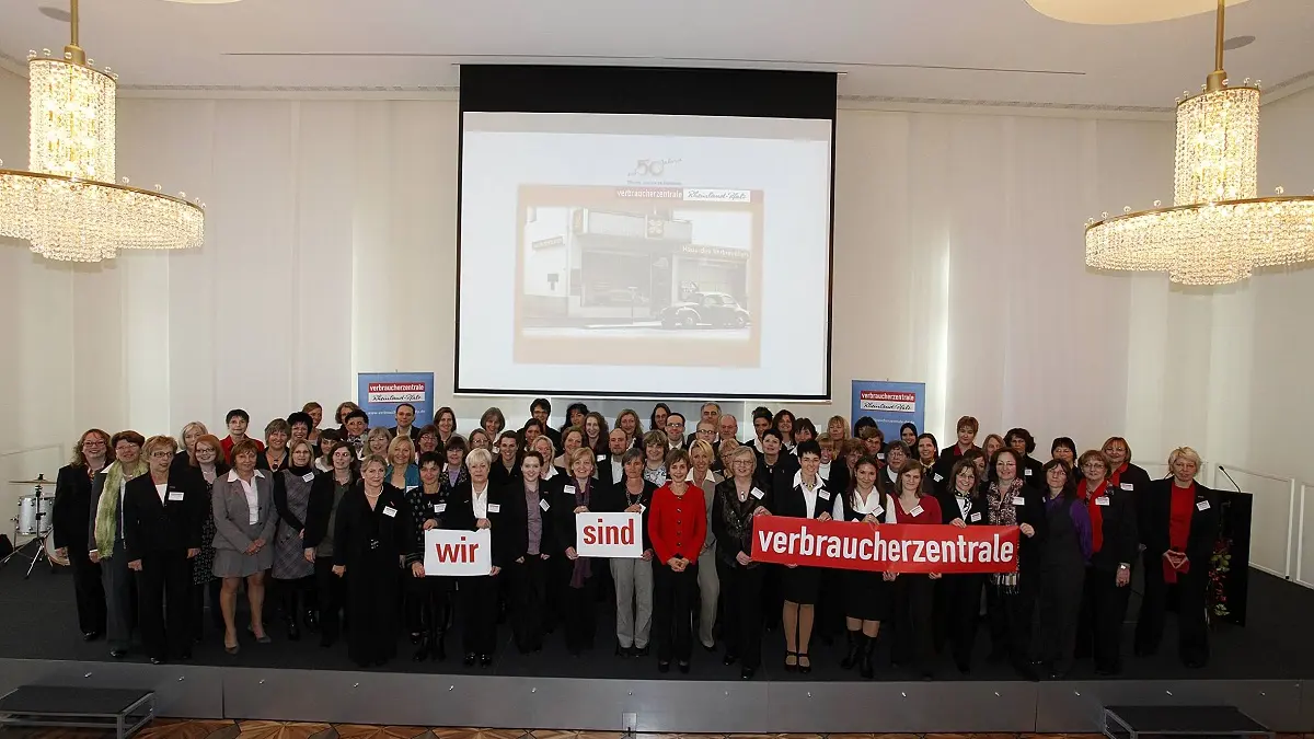 Viele Menschen auf eienr Bühne, die in die Kamera blicken und ein Schild vor sich halten.