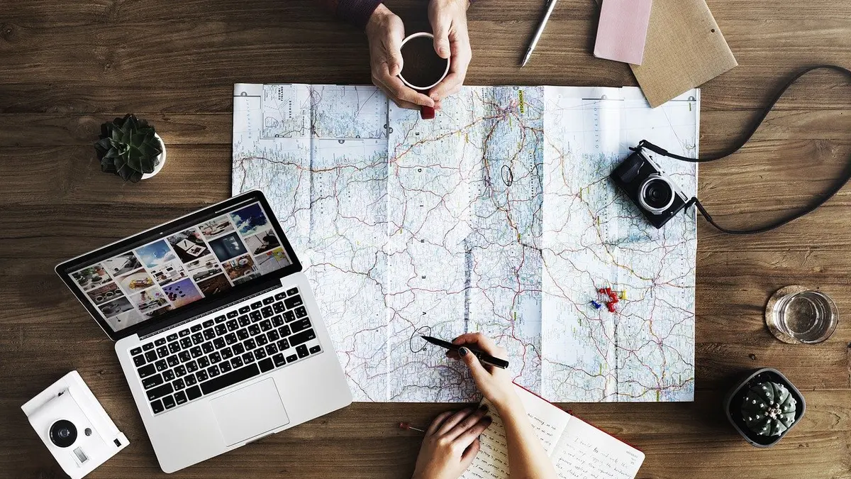 Zwei Personen planen eine Reise an einem Tisch mit Laptop, Weltkarte, Fotoapparat und weiteren Utensilien.
