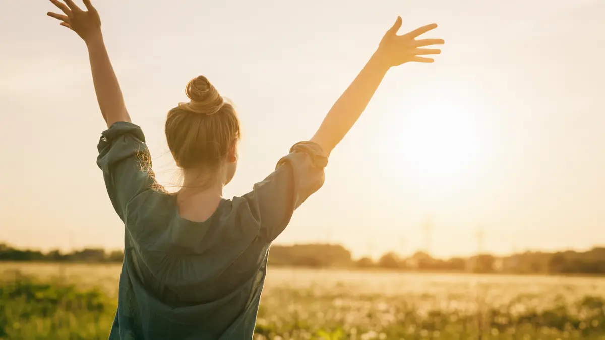 Eine junge Frau steht vor einem Getreidefeld in der Sonne und streckt die Arme in die Höhe.