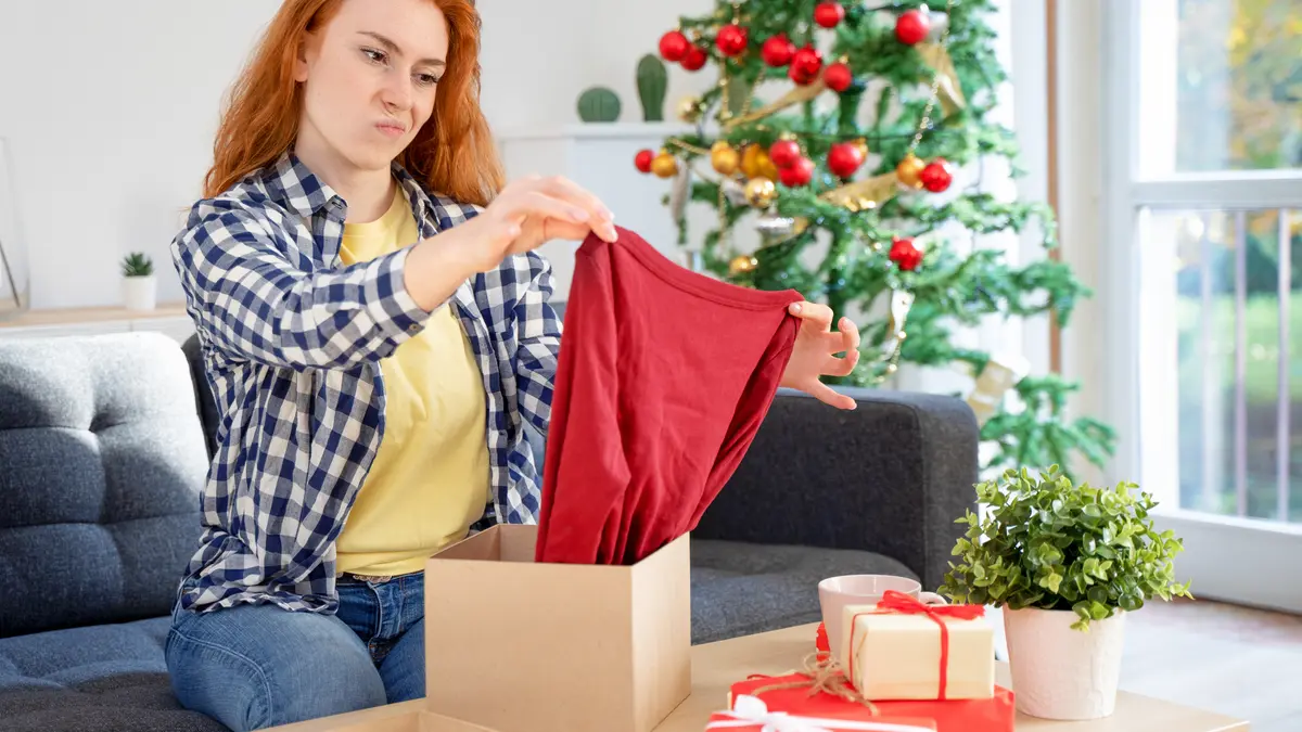 Frau sitzt vor einem Weihnachtsbaum auf der Couch und öffnet ein Geschenk, das ihr nicht gefällt