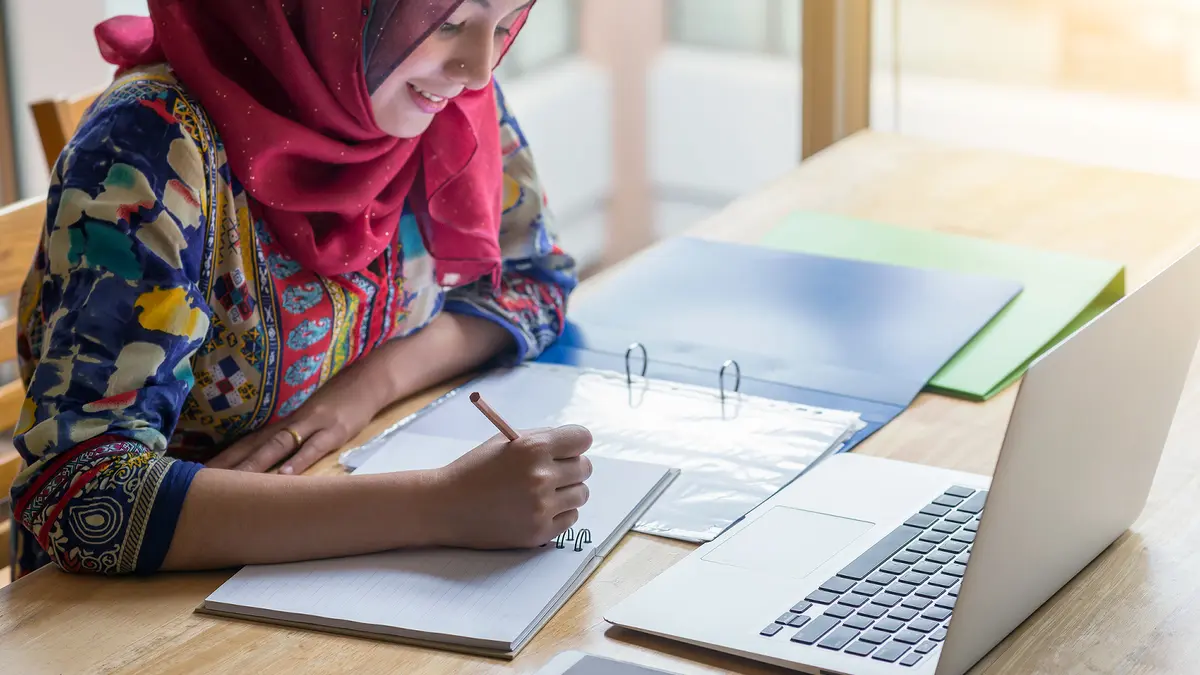Junge Frau mit pinkem Kopftuch sitzt mit Unterlagen vor ihrem Laptop