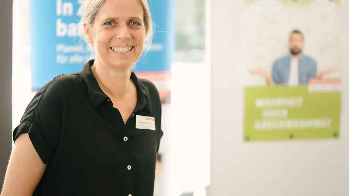 Eine Frau steht vor einem Messestand und lächelt in die Kamera
