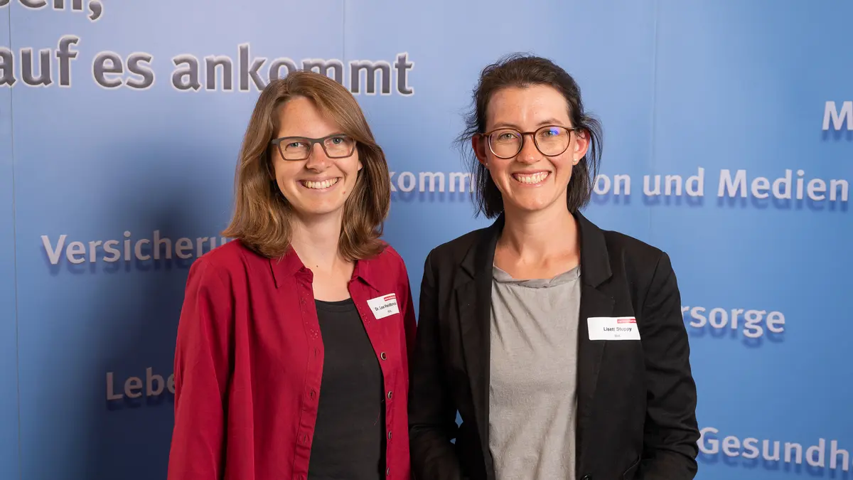 Zwei Frauen lächeln vor eienr Fotowand in die Kamera.