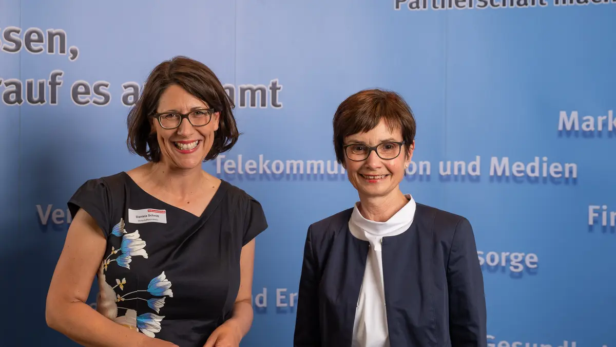 Zwei Frauen lächeln vor einer Fotowand in die Kamera.