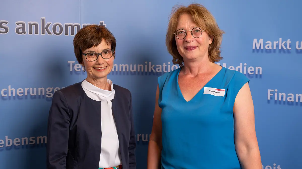 Zwei Frauen lächeln vor einer Fotowand in die Kamera.