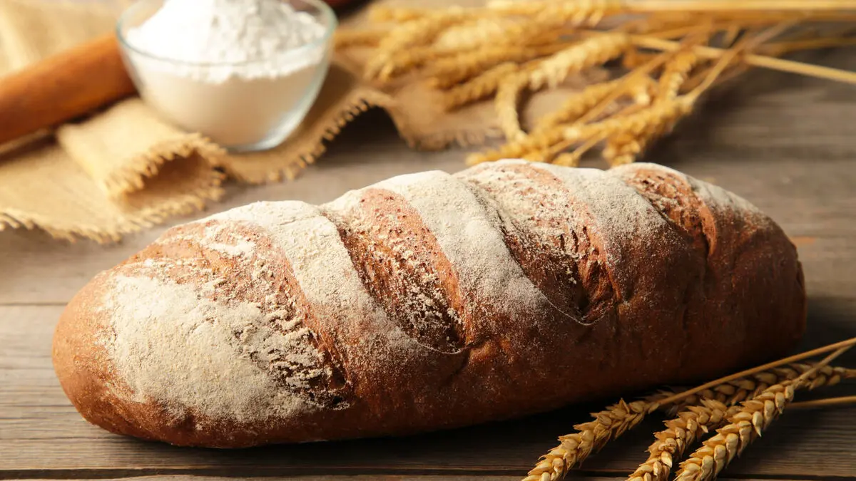 Ein länglicher Laib Brot liegt auf einem Holztisch, daneben liegen Ähren und einen kleine Schüssel Mehl steht auf dem Tisch.. 