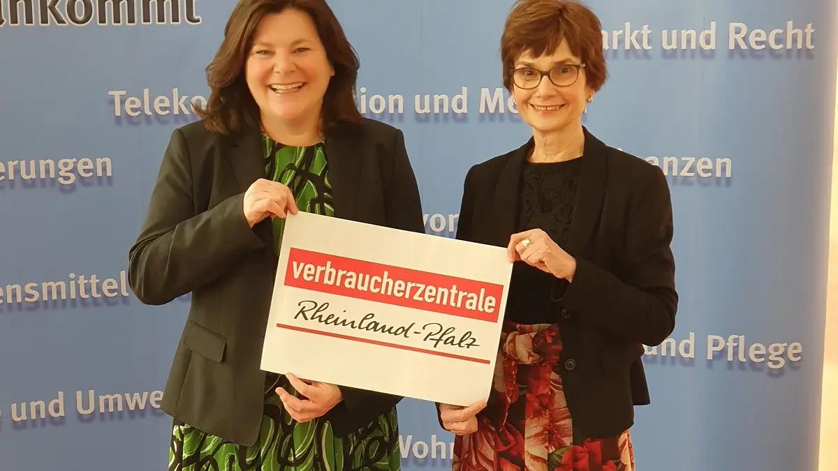 Zwei Frauen stehen vor einer baluen Wand und halten ein Schild mit Verbraucherzentrale Rheinland-Pfalz zwischen sich.