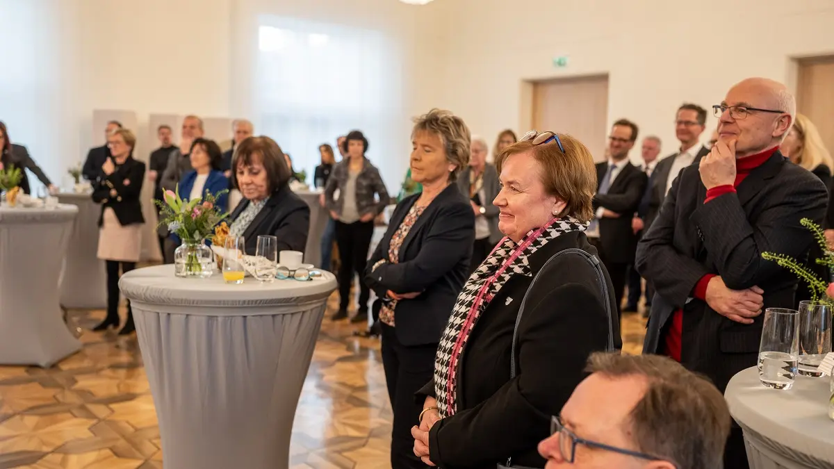 Einige Menschen stehen an Stehtischen vor einer Bühne und verfolgen das Geschehen auf der Bühne.