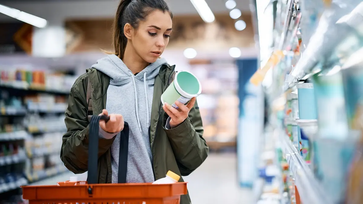 Eine Frau steht im Supermarkt und schaut kritisch auf eine Verpackung eines Lebensmittel.