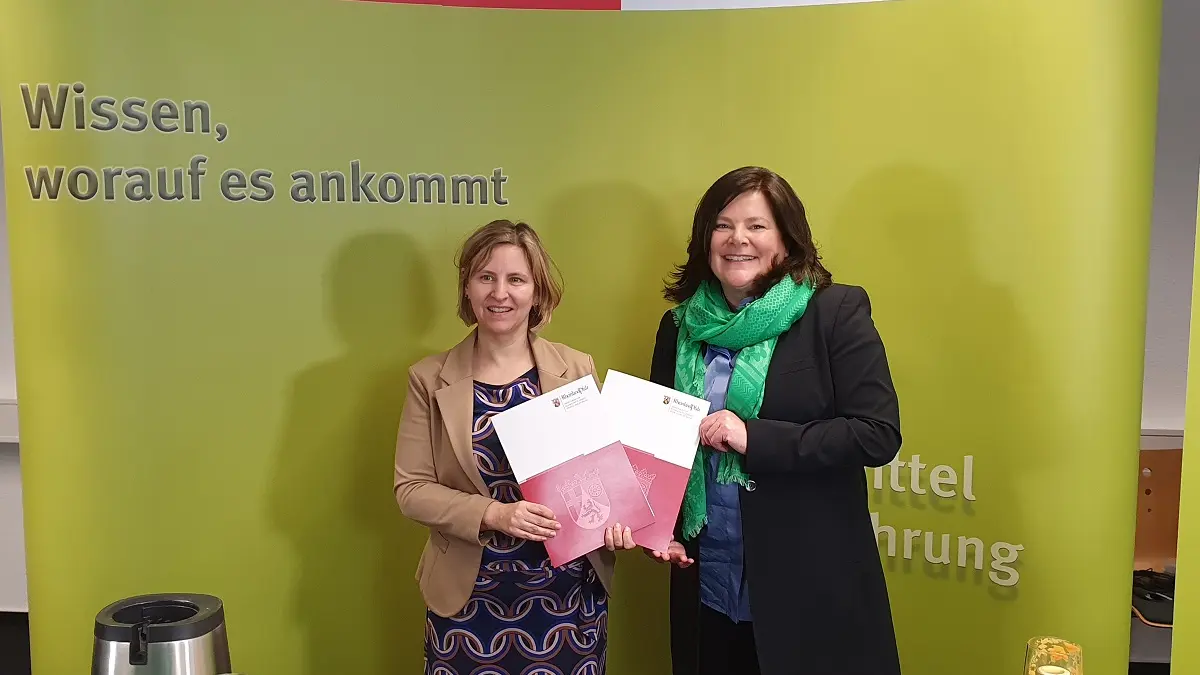 Zwei Frauen vor eienr grünen Wand. Sie halten zwei Dokumentenmappen in die Kamera.