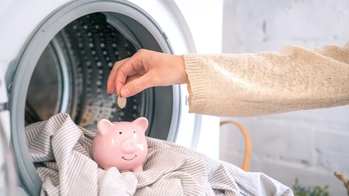 Eine Hand steckt ein Geldstück in ein Sparschwein, dass in einer Waschmaschine steht.