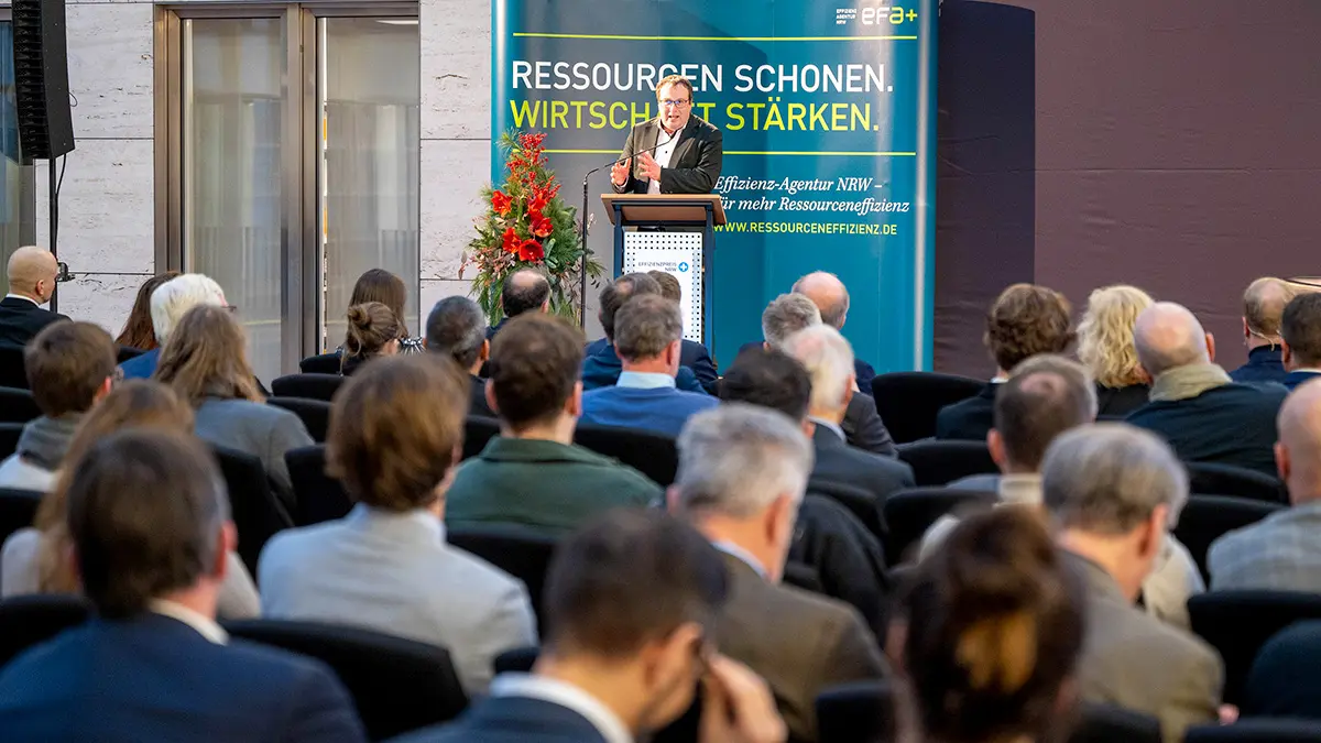 Ein Mann steht in der Ferne am Rednerpult, im Vordergrund sitzen zahlreiche Gäste.
