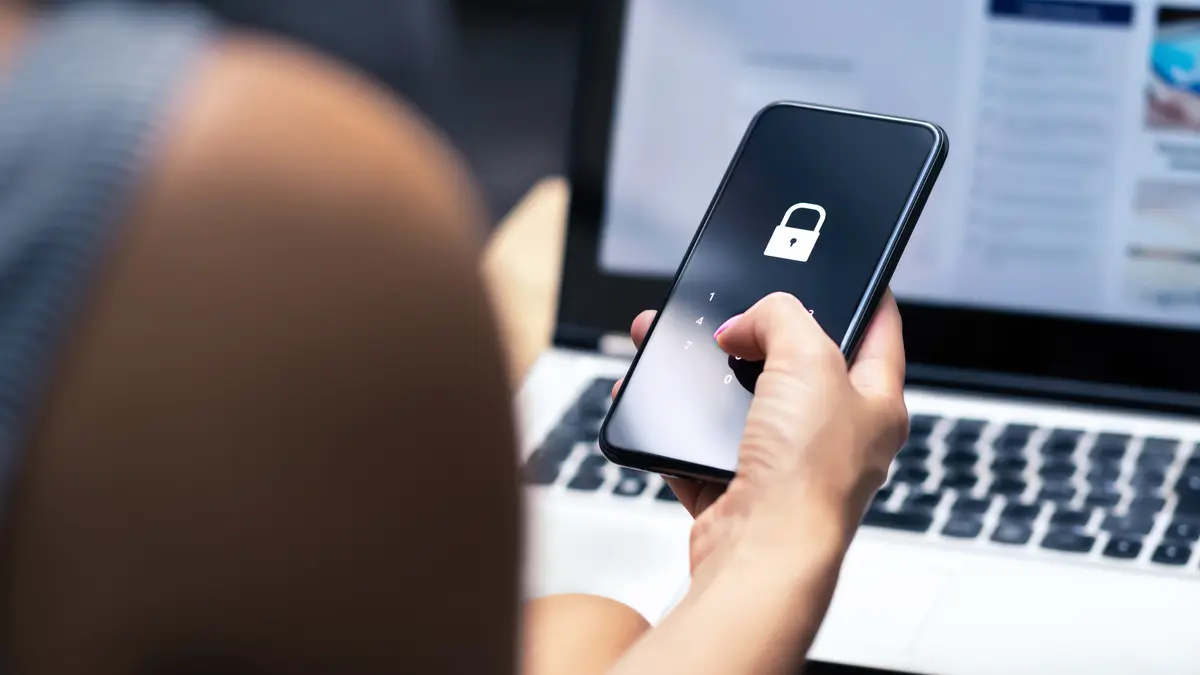 Eine Person sitzt an einem Laptop und hält ein Smartphone in der Hand. Auf dem Smartphone ist ein Verschlüsselungssymbol.