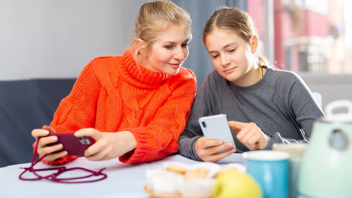 Mutter sitzt mit Tochter am Tisch und schaut auf ihr Smartphone.