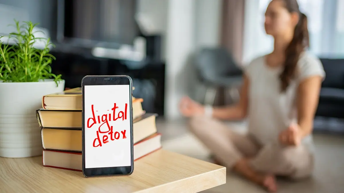 Im Vordergrund liegt ein Handy mit den Wörtern: digital detox auf dem Bildschirm. Im Hintergrund macht eine Frau Yoga