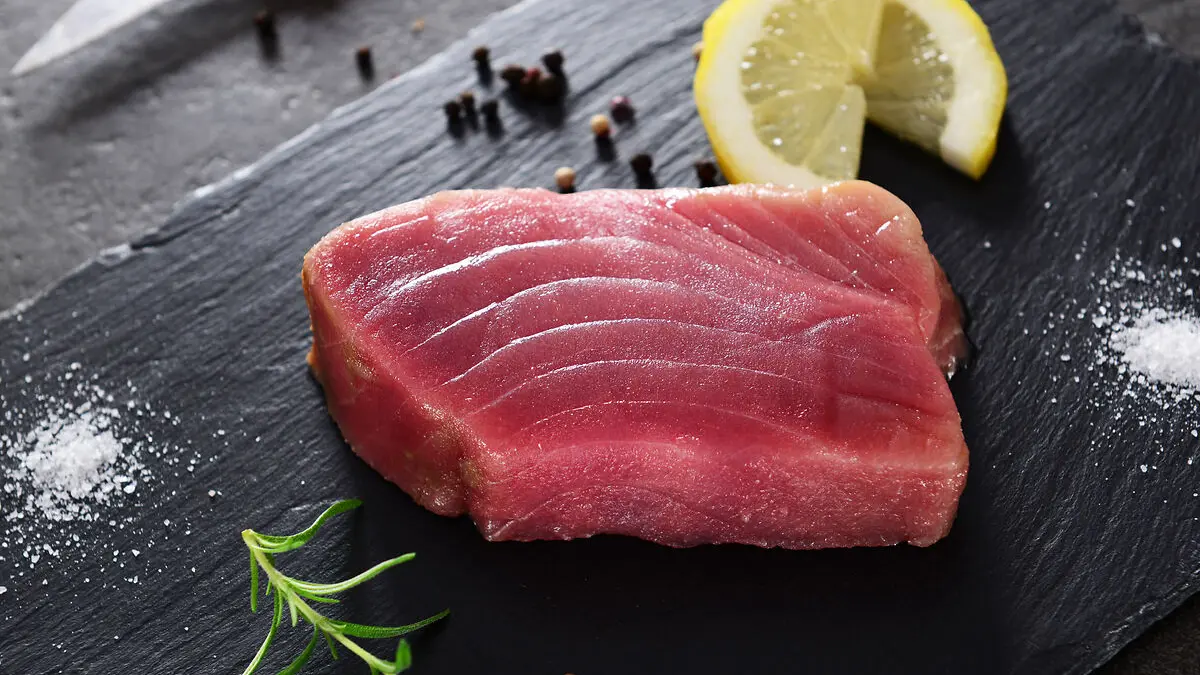 Ein rohes Stück Thunfisch liegt auf einem schwarzen Brett, daneben eine Zitronenscheibe, schwarze Pfefferkörnern und ein Zweig Rosmarin.