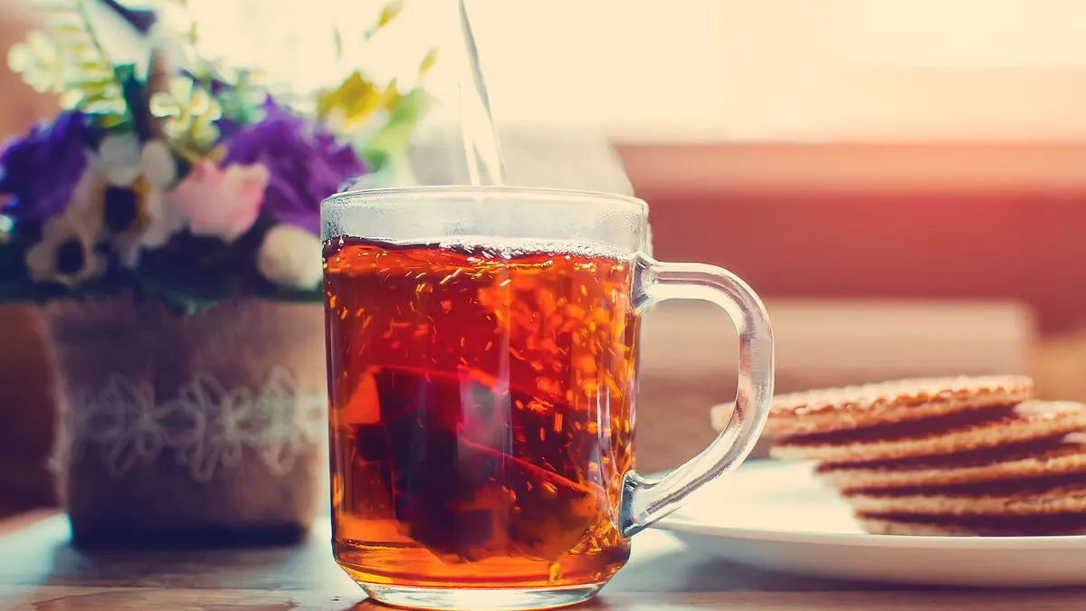 Eine Tasse Tee steht auf einem Tisch, links davon stehen Blumen, rechts davon ein Teller mit Keksen