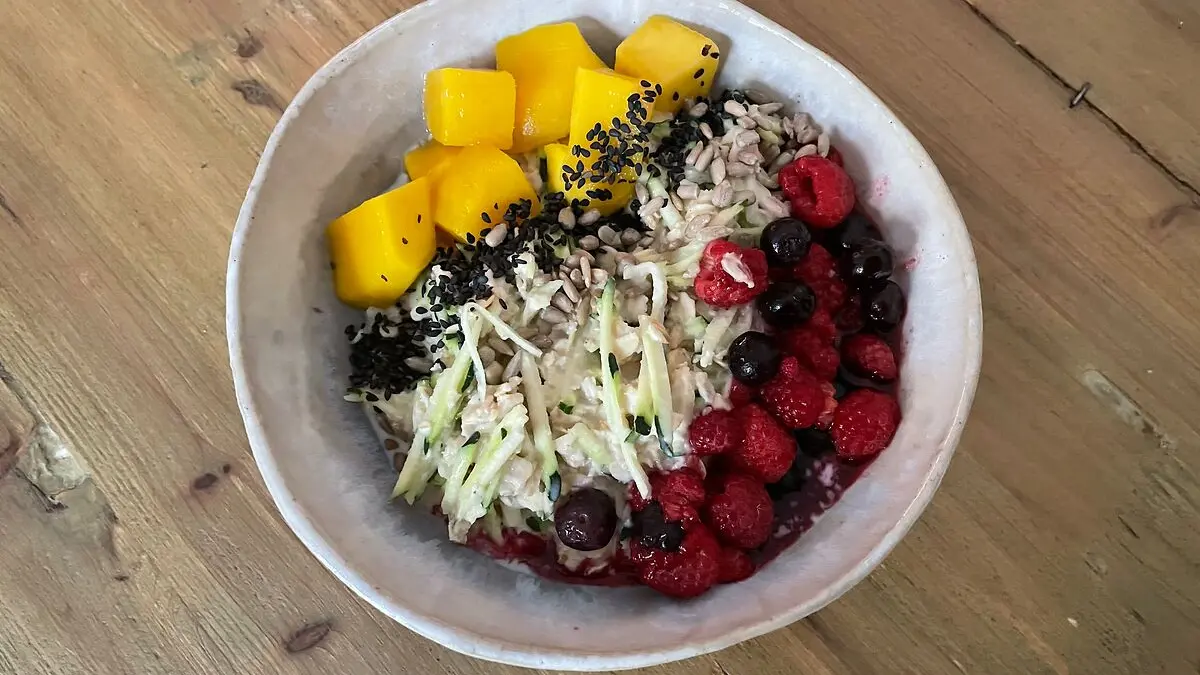 Ein Schüssel mit Haferbrei steht auf einem Tisch. Garniert ist der Brei mit Mango, Beeren und Zucchini.