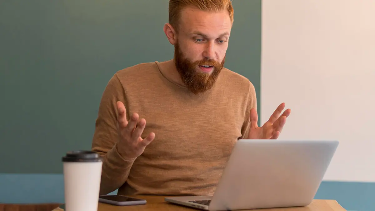 Ein Mann sitzt vor einem Laptop und hebt verärgert die Hände
