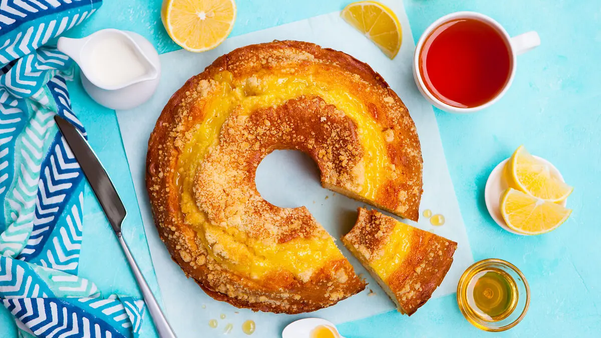 Ein Rührkuchen von oben, daneben befinden sich eine aufgeschnittene Zitrone, eine Tasse Tee, ein Kännchen Milch, ein Messer und ein Löffel.