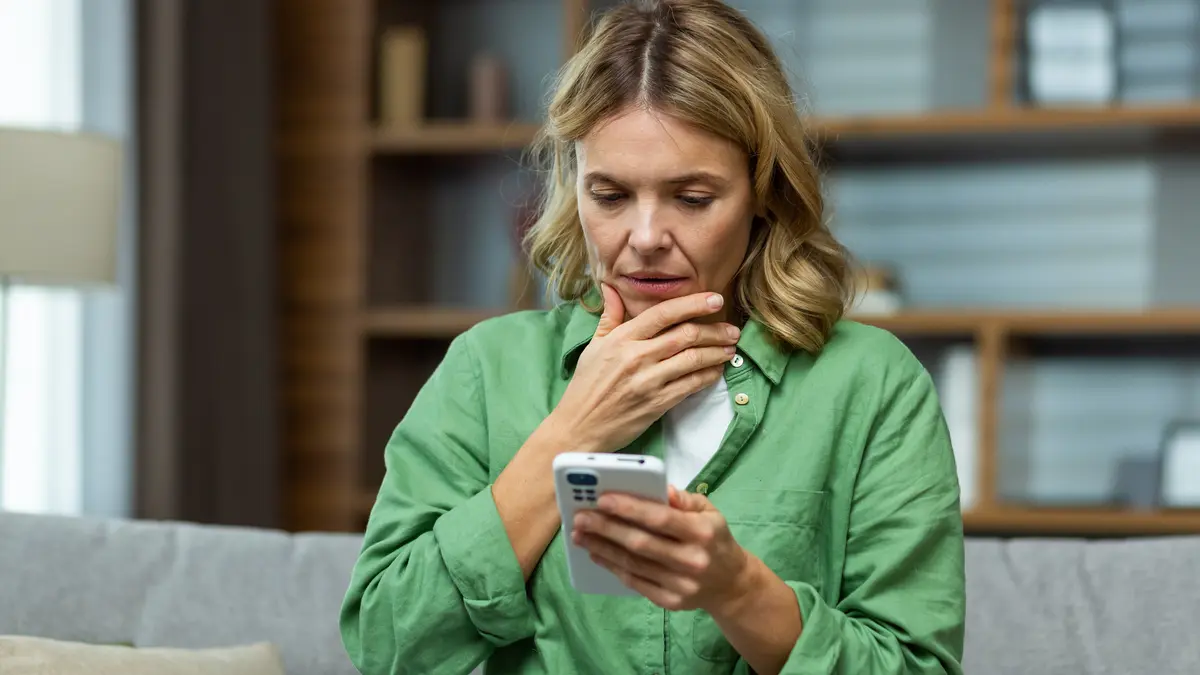 Eine Frau schaut fragend/ erschrockend auf ein Smartphone in ihrer Hand. Die andere Hand hält sie an ihr Kinn.