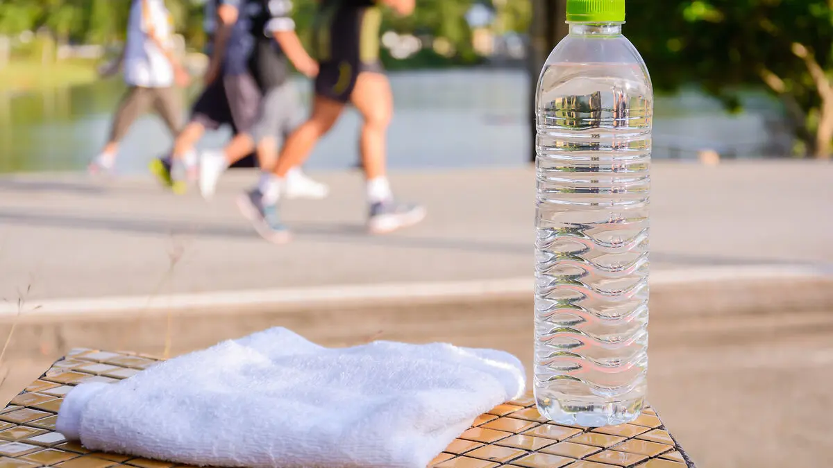 Im Vordergrund steht eine Wasserfasche aus PET, daneben liegt ein weißes Handtuch. Im Hintergrund joggt eine kleine Gruppe Läufer.