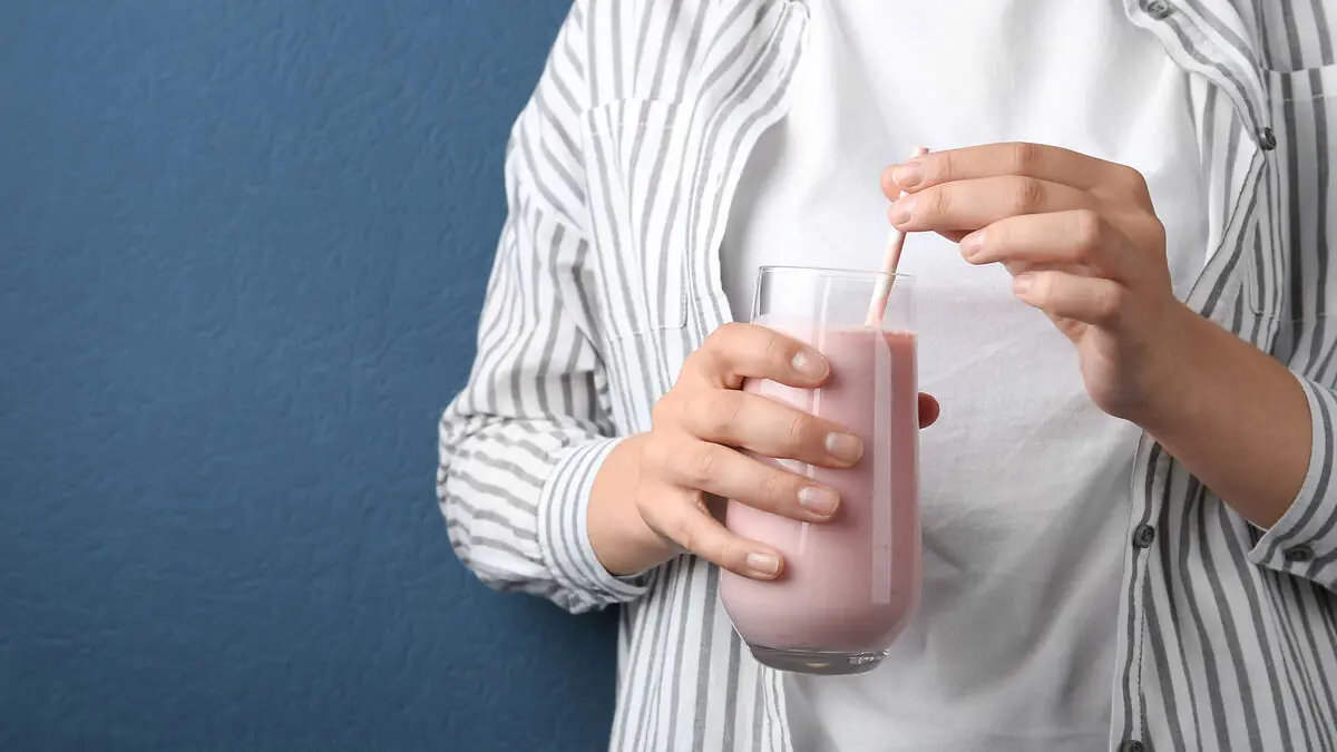 Eine Hand hält ein Glas mit einer hellrosa Trinkmahlzeit, drei Finger der anderen Hand umschließen den Strohhalm, der im Getränk steckt.
