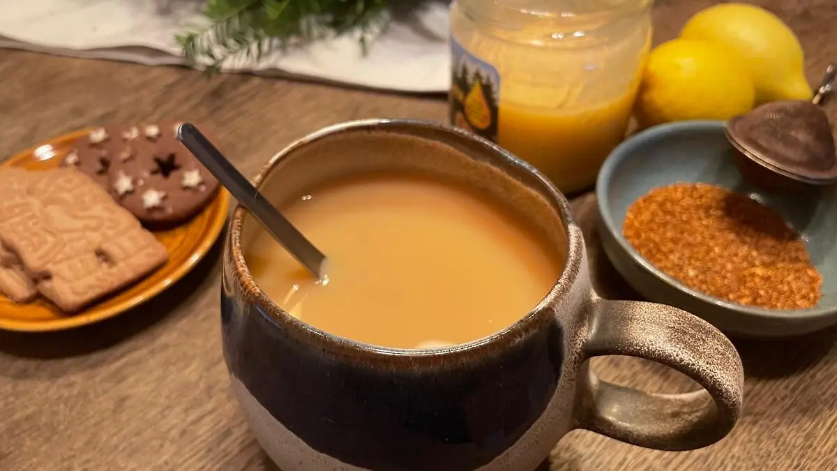Eine Tasse hellbrauner Tee mit Löffel steht auf einem Holztisch, umgeben von Honig, Zitronen, losen Teeblättern und einem Teller mit Weihnachtsgebäck.