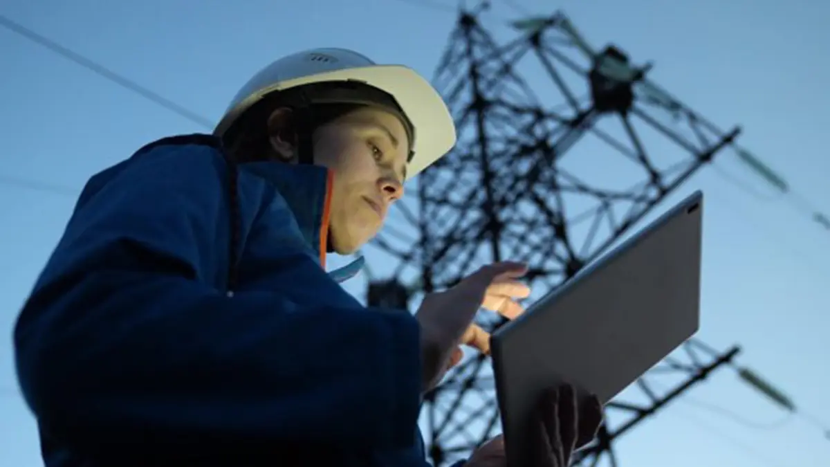 Person mit Schutzhelm nutzt ein Tablet vor einem Strommast, vermutlich zur Überprüfung oder Wartung von Energieinfrastruktur.