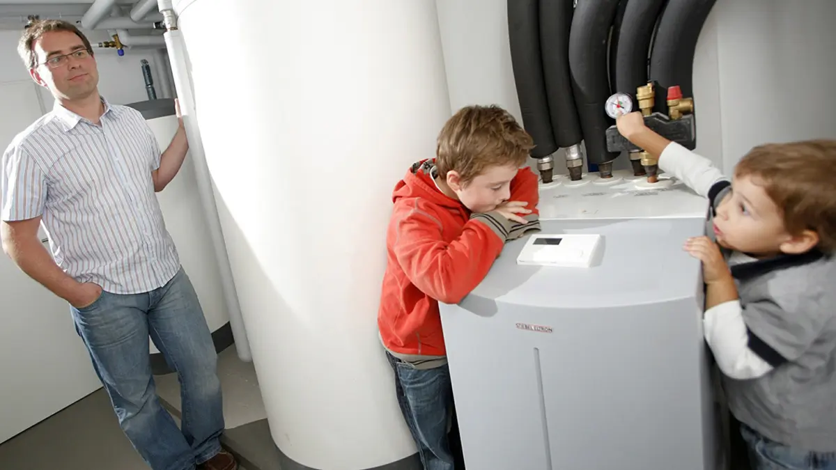 Mann und zwei Jungen sind im Heizungskeller und schauen sich die Wärmepumpe und den Wassertank an