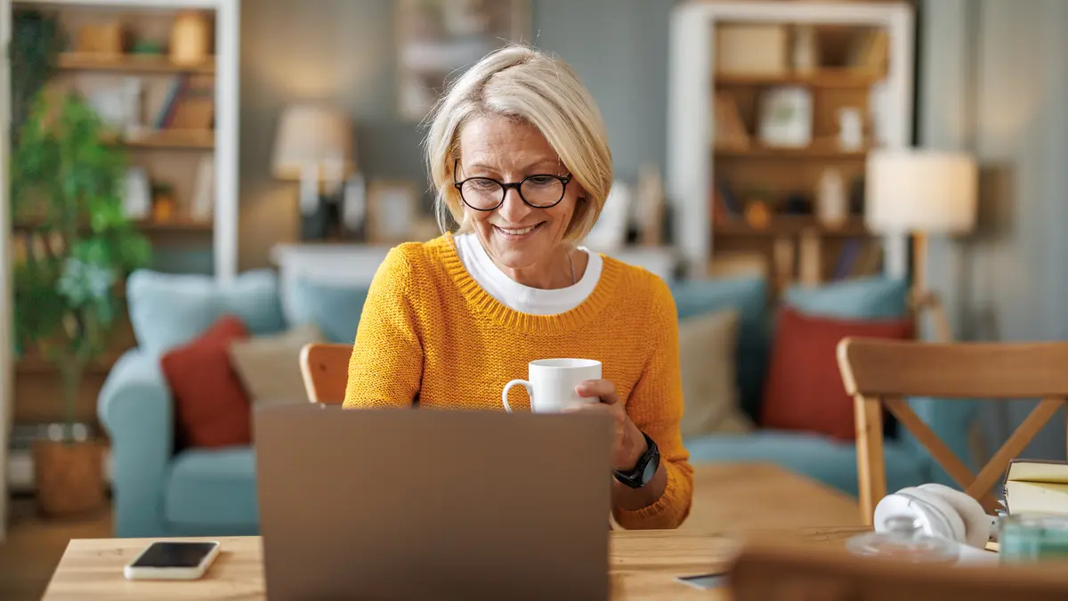 Eine blonde Frau mittleren Alters sitzt am Esstisch vor ihrem Laptop im Wohnzimmer. Sie hält eine weiße Tasse in ihrer Hand, schaut auf das Display und lächelt.