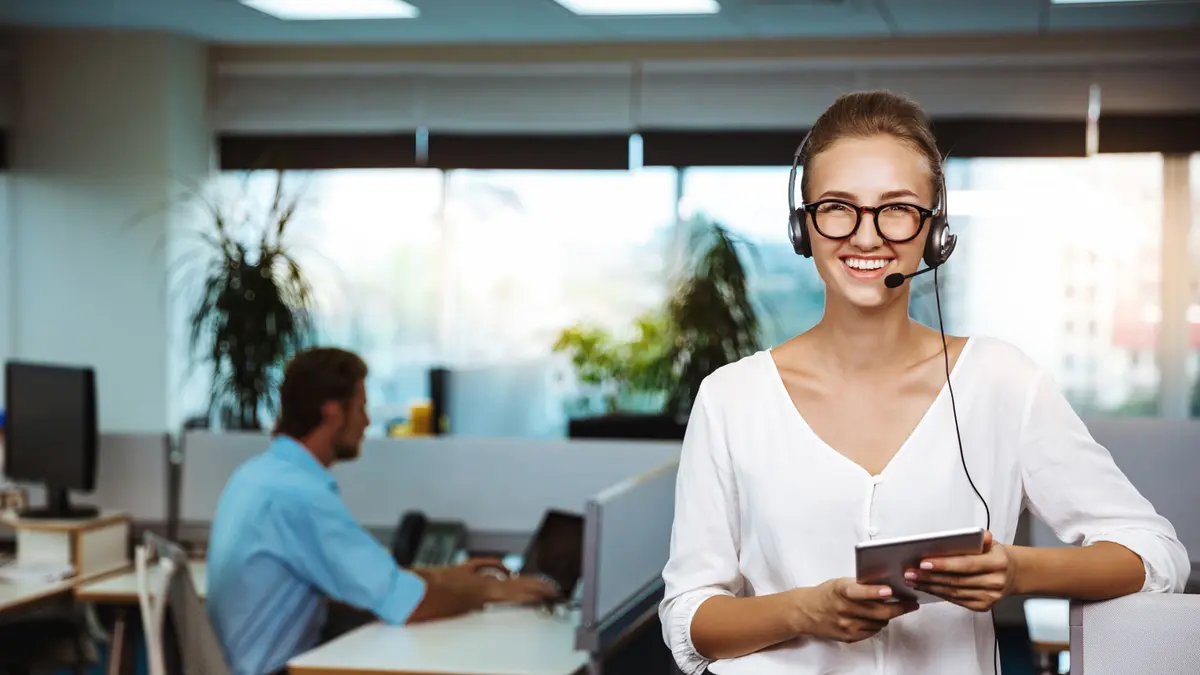 Eine Frau mit Headset steht in einem Büro, hinter ihr sitzt ein Mann am PC