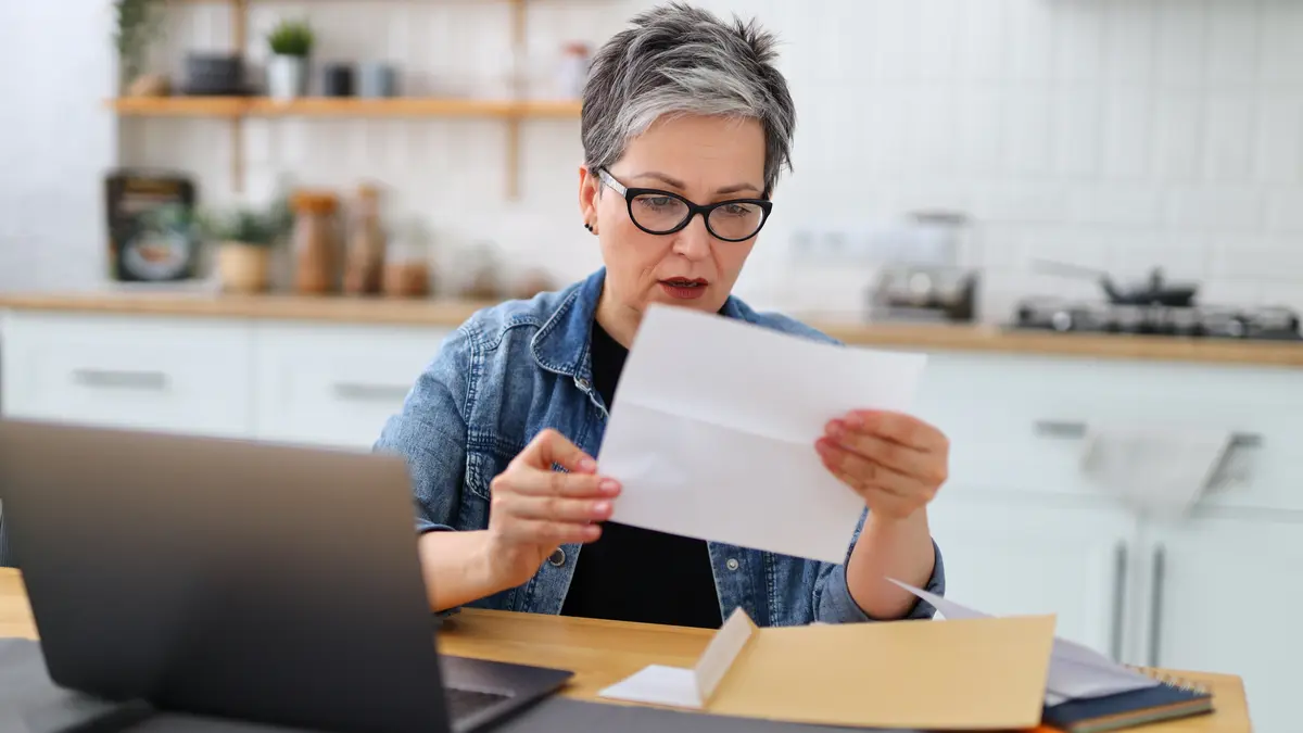 Eine Frau sitzt an einem Tisch mit Laptop und blickt erschrocken auf einen Brief.