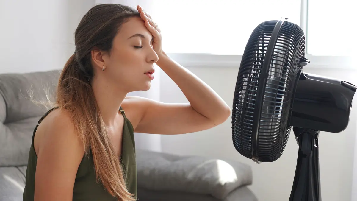 Erschöpfte Frau sitzt bei Hitze vor einem Ventilator im Wohnzimmer.