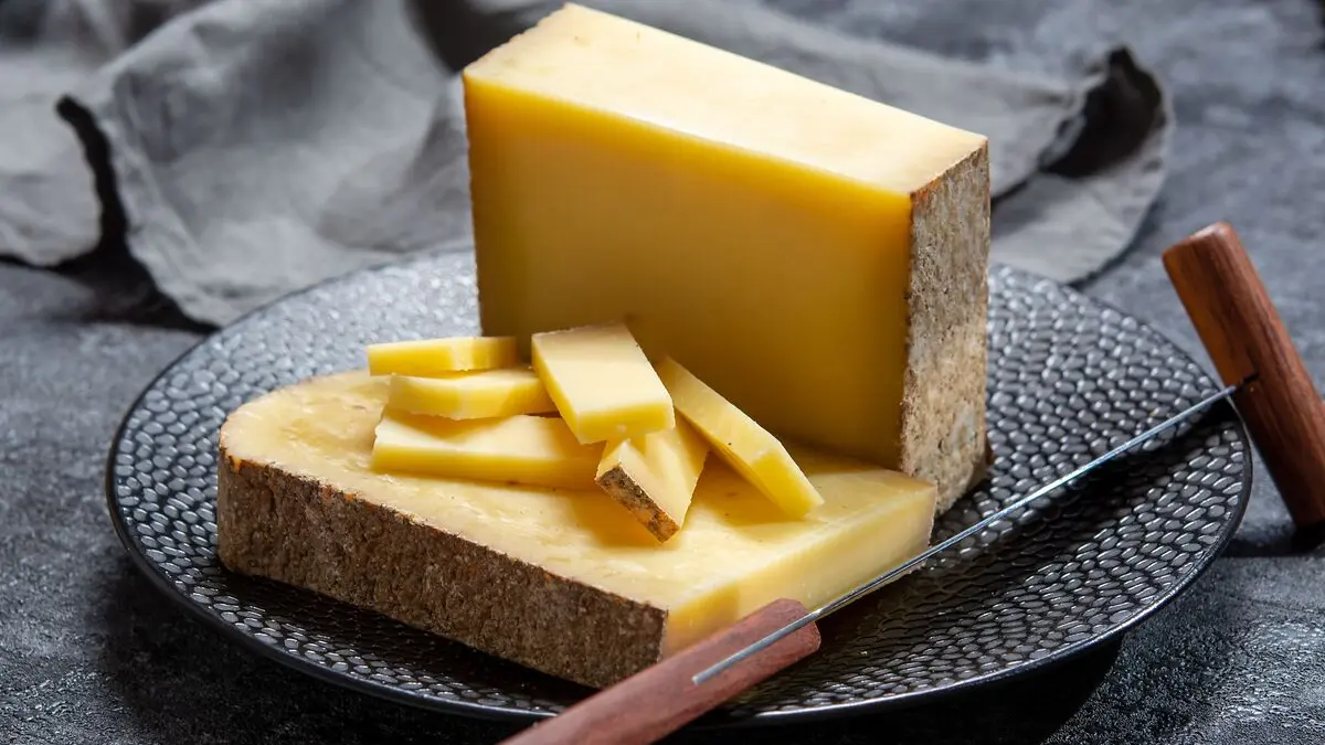 Zwei große Stücke und mehrere kleine Stücke Bergkäse liegen auf einem schwarzen Teller, daneben ein Käsemesser