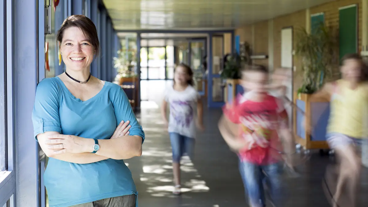 Eine Lehrkraft steht im Flur eines Schulgebäudes, Kinder rennen vorbei