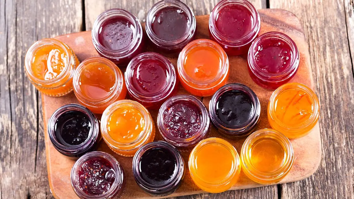 Verschiedene geöffnete Gläser mit Marmelade stehen auf einem Tablett. Man blickt von oben auf die geöffneten Gläser und sieht gelbe, orangefarbene, rote und dunkelrote Marmeladen.