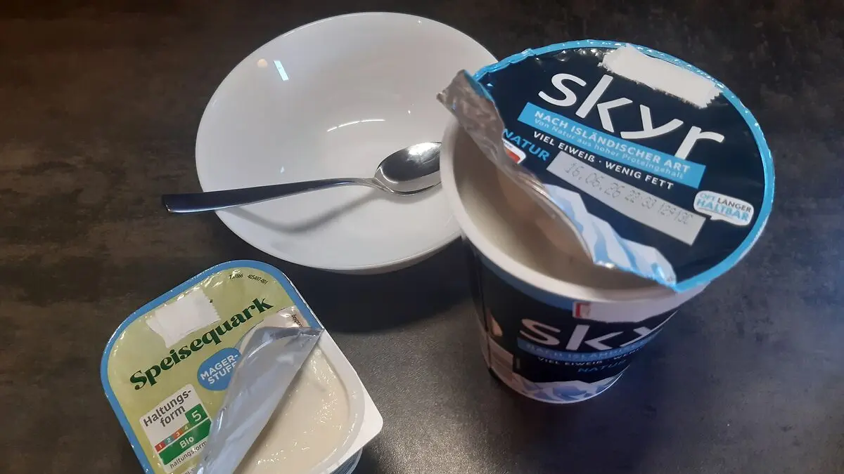 Eine Packung Quark und ein Becher Skyr stehen auf einem Tisch neben einer weißen Schüssel, in der ein kleiner Löffel liegt.