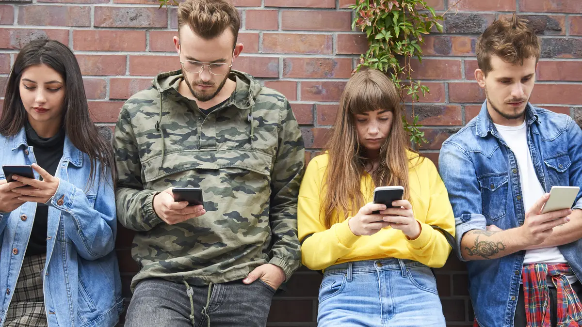 Vier junge Erwachsene stehen an eine Mauer gelehnt und schauen in ihr Handy.