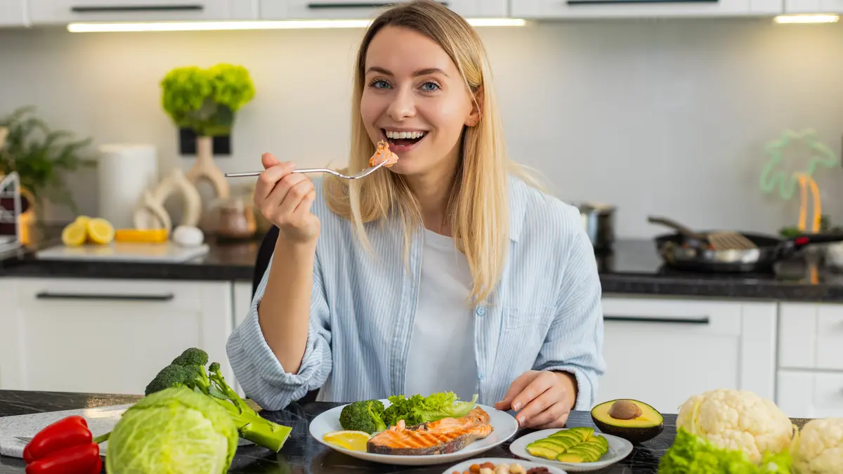 Eine Frau steht in der Küche und lacht in die Kamera. Vor ihr liegt ein Teller mit einem gebratenen Stück Lachsfilet. Ein Stück führt sie mit der Gabel zu ihrem Mund.