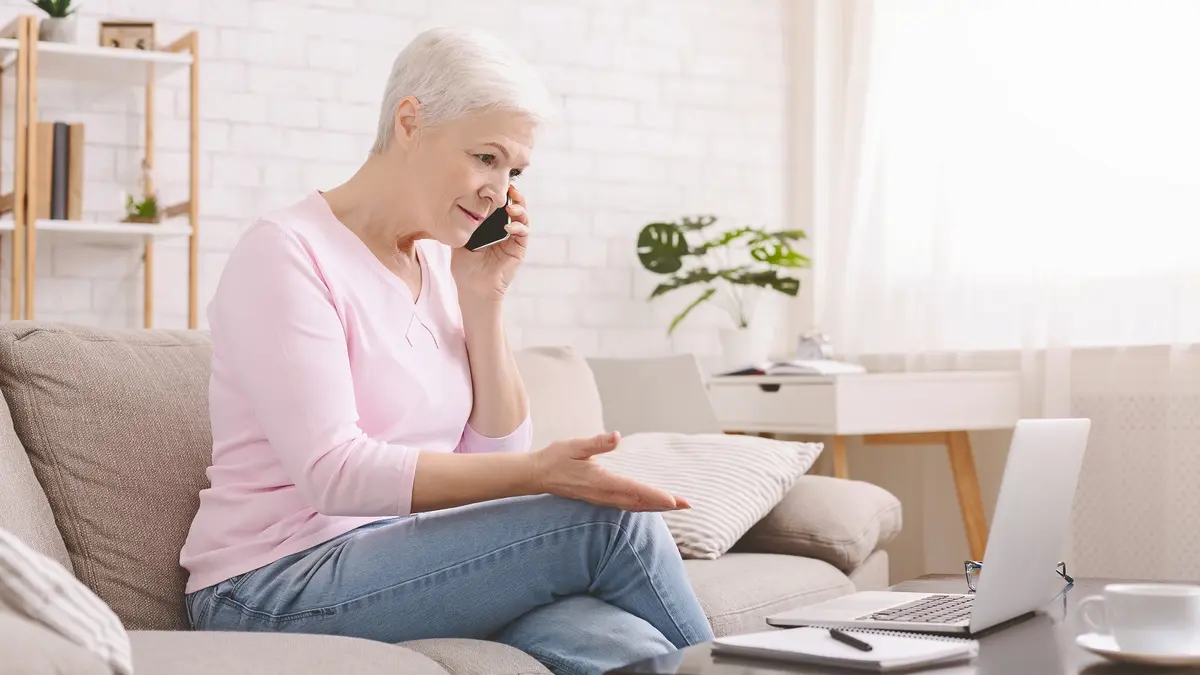 Eine ältere Frau sitzt zu Hause auf dem Sofa und telefoniert mit dem Smartphone. Vor ihr steht ein geöffneter Laptop auf dem Couchtisch. Mit der freien Hand gestikuliert sie und wirkt besorgt oder ratlos.