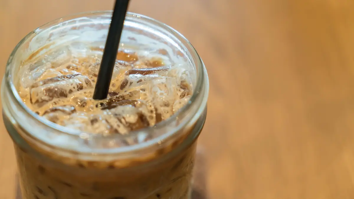 Ein Glas mit Kaffee und Eiswürfeln auf einer Holzoberfläche. In der Mitte steckt ein Strohhalm.