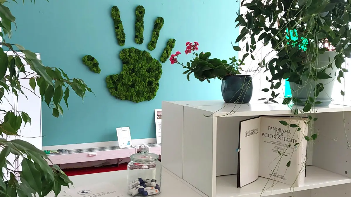 Blick durch weiße Regale und Pflanzen auf eine türkisfarbene Wand, an der aus Moos eine Hand geformt ist.