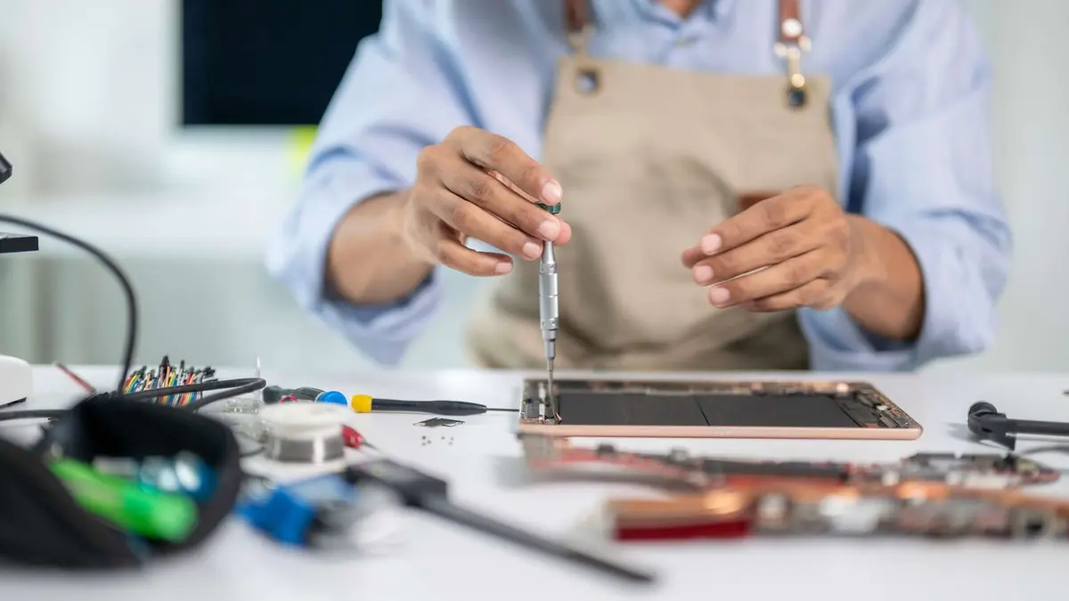 Ein Techniker repariert mit einem kleinen Schraubenzieher ein geöffnetes Tablet an einem Werkstatttisch voller Werkzeug.