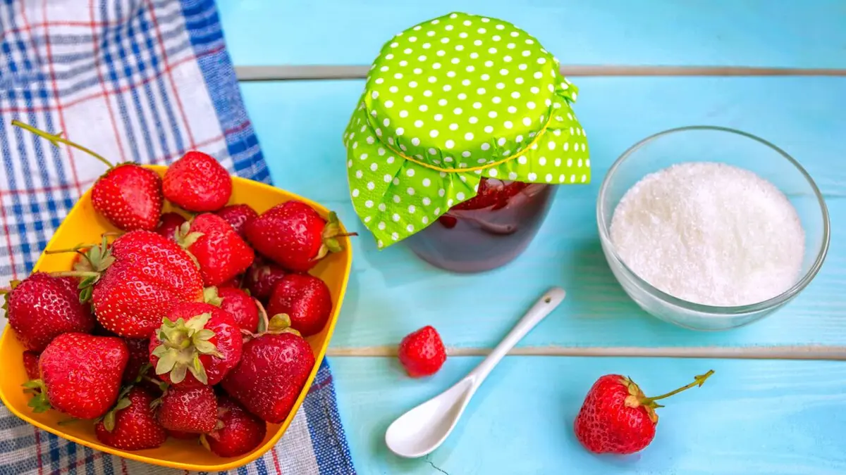 Auf einem Tisch stehen eine Schüssel mit Erdbeeren, ein Glas Marmeladen und eine Schüssel mit Gelierzucker.