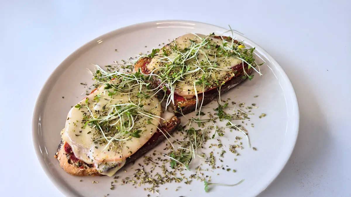 Auf einem Teller liegen zwei überbackene Brotscheiben mit geschmolzenem Käse und frischen Sprossen, bestreut mit Kräutern. Das Gericht wirkt schlicht, aber fein angerichtet und erinnert an ein kreatives warmes Brotgericht oder eine schnelle Mahlzeit.