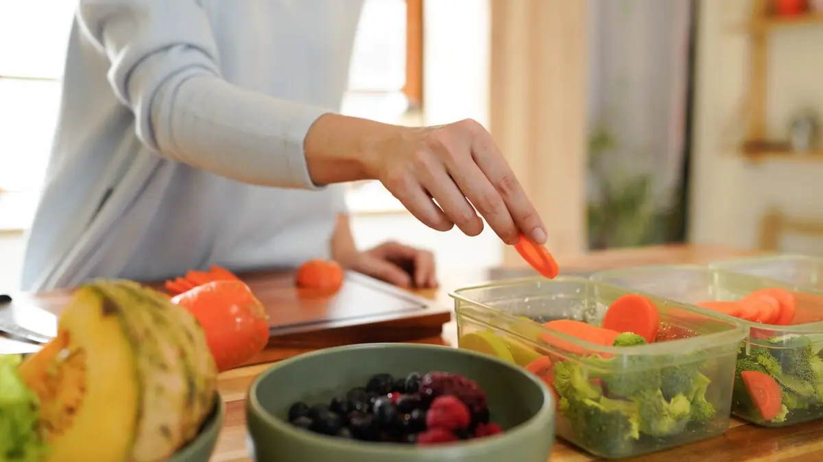 Eine Frau gibt Karottenstückchen in Meal Prep-Glasbehälter, in denen bereits anderes Gemüse liegt.