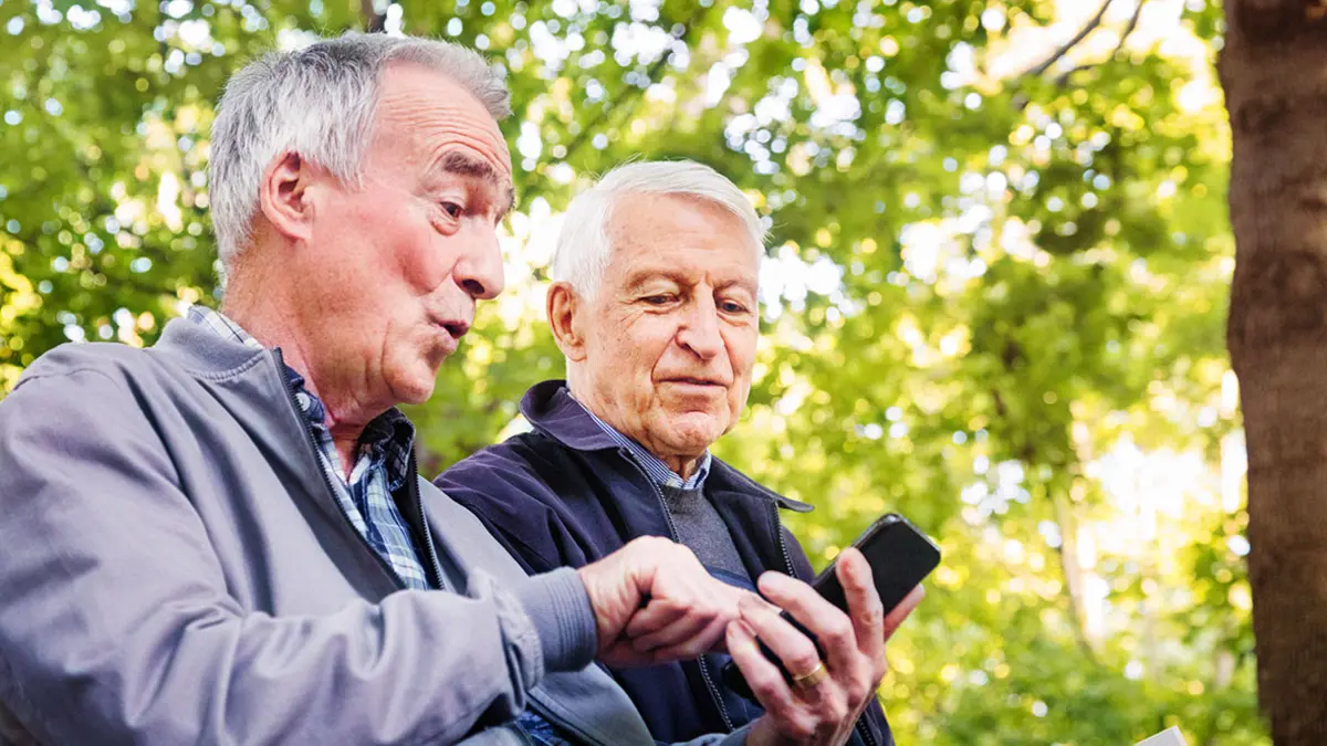 Zwei ältere Männer sitzen auf einer Bank im Park, einer zeigt dem anderen etwas auf seinem Smartphone.