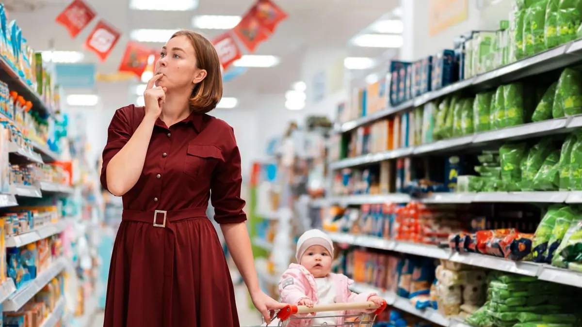 Eine Frau in dunkelrotem Kleid steht nachdenklich im Supermarktgang, den Finger an den Lippen, und blickt zu den Regalen hoch. Vor ihr schiebt sie einen Einkaufswagen, in dem ein Baby mit rosa Jacke und weißer Mütze sitzt."